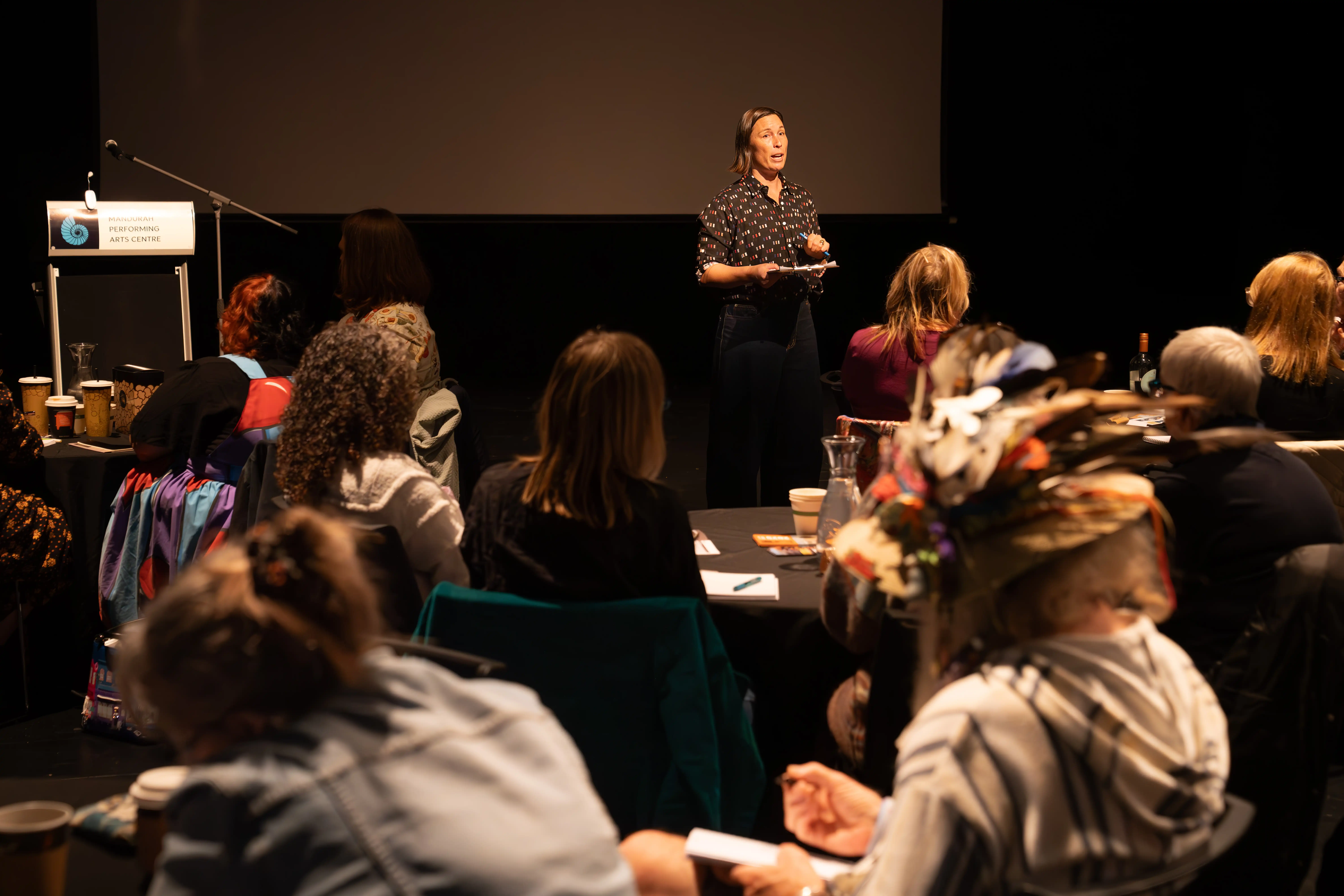 Presenter standing at the front of a room addressing an audience seated at tables.